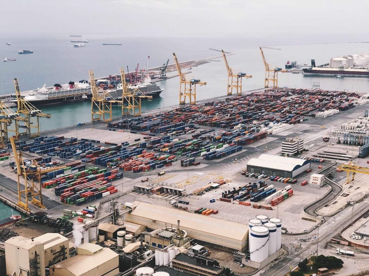 Expansive aerial view of a bustling container port with large cranes and cargo ships under a cloudy sky.